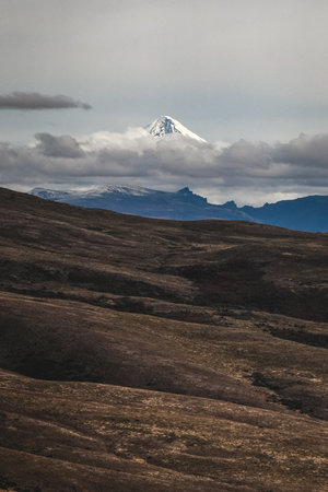 Distant snow-covered mountain peak of Lanin volcano rises above a vast, undulating Patagonian landscape under a cloudy sky. Neuquen, Argentinaの写真素材