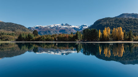 Lake Moquehue reflecting colorful trees line and snowcapped mountains in the background under a clear blue sky. Neuquen, Argentinaの写真素材