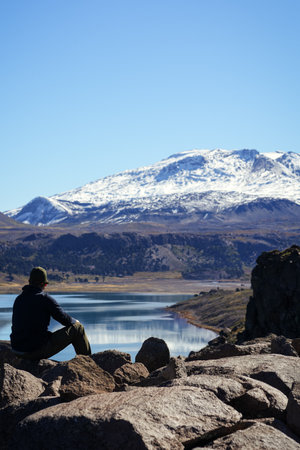 Unrecognizable man sitting on rock looking at Lake Caviahue and snowcapped Copahue volcano in the background under a clear blue sky. Neuquen, Argentinaの写真素材