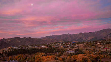 Scenic view of Andacollo village nestled among autumn trees and mountains under a vibrant pink and purple dusk sky with the moon. Neuquen, Argentinaの写真素材