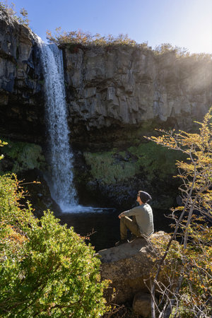 Man sits peacefully on a rock, gazing at the flowing Cascada de la Vieja Usina waterfall under autumn sunlight. Caviahue, Neuquen, Argentinaの写真素材