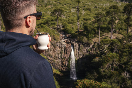 Caucasian man drinks mate, overlooking Cascada Coloco waterfall surrounded by lush greenery and Araucaria Araucana trees in Neuquen, Patagonia, Argentinaの写真素材