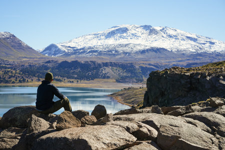 Man sitting on rocks overlooking the serene Lago Caviahue and snowcapped Copahue volcano in the background under a clear blue sky. Neuquen, Argentinaの写真素材