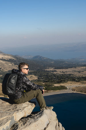 Man with a backpack sits on a rock at Batea Mahuida, enjoying the scenic view of the lake and landscape. Villa Pehuenia, Neuquen, Argentinaの写真素材
