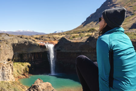 Woman in a beanie and sunglasses sits on a rocky edge overlooking the Salto del Agrio waterfall. Neuquen, Argentinaの写真素材