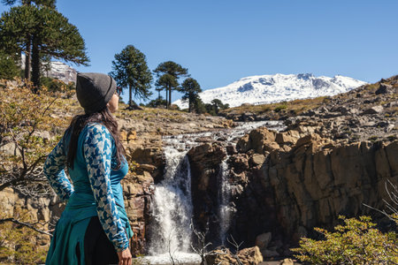 Woman in beanie looks at Cascada del Gigante waterfall surrounded by Araucaria trees, with snowcapped Copahue volcano in the background. Caviahue, Neuquen, Argentinaの写真素材
