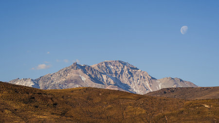 Snow-dusted Domuyo volcano peak under a clear blue sky with a visible moon. Rugged, brown hills in the foreground. Neuquen, Argentinaの写真素材