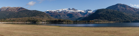 Panoramic view captures serene lake Moquehue, framed by lush trees and majestic snow dusted mountains. Neuquen, Argentinaの写真素材