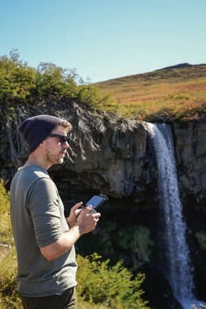 Man stands on a rock outcropping, holding drone controller, with Cascada de la Usina waterfall in the background. Caviahue, Neuquen, Argentinaの写真素材