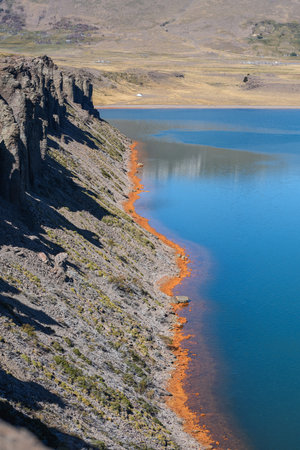 Rocky shoreline with orange mineral deposits along the water's edge of Lake Caviahue with blue waters. Neuquen, Patagonia, Argentinaの写真素材