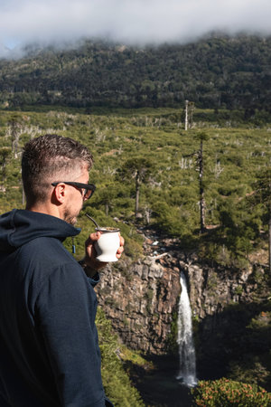 Man enjoys mate drink while overlooking Cascada Coloco waterfall surrounded by lush vegetation and Araucaria trees in Neuquen, Patagonia, Argentinaの写真素材