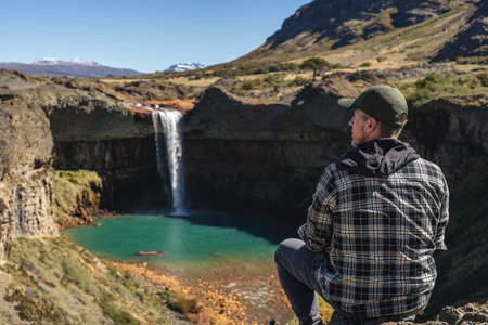 Man tourist gazing at the Salto del Agrio waterfall plunging into a turquoise pool, with snow-capped mountains in the distance. Neuquen, Argentinaの写真素材