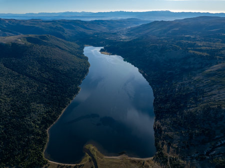 Aerial view of Rucachoroi lake surrounded by forested hills with araucaria trees under a clear sky and distant mountain ranges visible in the distance. Neuquen, Argentinaの写真素材