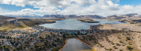 Panoramic aerial view of Caviahue village nestled beside lake, surrounded by mountains with patches of snow under cloudy sky. Neuquen, Argentinaの写真素材