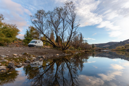 White van is parked by a calm river reflecting trees and a cloudy sky, creating a serene landscape. Alumine, Argentinaの写真素材