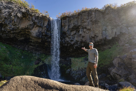 Happy man stands on a rock pointing towards a tall waterfall cascading down a dark rocky cliff. Caviahue, Neuquen, Argentinaの写真素材