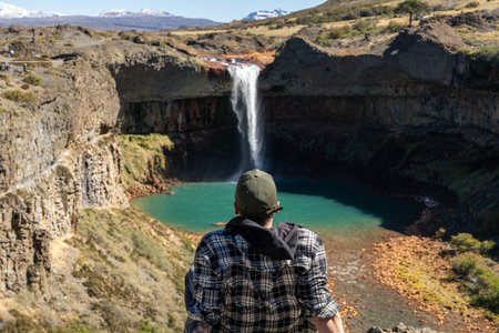 Man back view in a plaid shirt and cap stands on a rocky edge overlooking the Salto del Agrio waterfall. Neuquen, Argentinaの写真素材