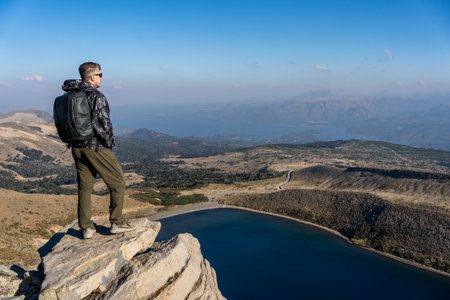 Man with a backpack stands on a rocky mountain top at Batea Mahuida, overlooking a lake and a vast landscape. Villa Pehuenia, Neuquen, Argentinaの写真素材