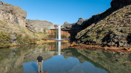 Man stands at the base of the Salto del Agrio waterfall, with the river and rocky cliffs surrounding it under a clear sky. Neuquen, Patagonia Argentinaの写真素材