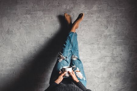 Girl in jeans on a gray background with coffee. Reads a book with his legs to the wall. Education, development.の写真素材