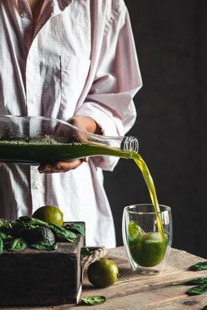 Woman pouring freshly blended green and white smoothie in glass jar with ingredients around on a rustic wooden table. Healthy eating and lifestyle concept. Detox diet. Recipe ideas.の写真素材