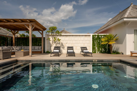 This captivating image showcases a luxurious swimming pool area at a tropical villa resort The scene exudes relaxation and sophistication with three elegant loungers arranged perfectly for sunbathing or quiet contemplation The wooden deck and pergola offer a touch of rustic charm enhancing the tranquil atmosphere The pools pristine water reflects the cloudy sky creating a serene visual appeal This image epitomizes the ultimate vacation lifestyle offering a glimpse into a world of comfort and luxury perfect for travel and lifestyle magazines or advertisementsの素材