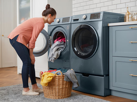 A young woman bends over in a bright modern laundry room while loading clothes into a front load washing machine The laundry room features shaker style cabinets and a woven laundry basket overflowing with colorful clothes ready to be washed The modern front load washing machine is a dark grey and sits next to the dryer The woman is wearing jeans and a pink blouse and her hair is in a neat bun The scene represents a clean and organized approach to household chores in a stylish and functional spaceの素材