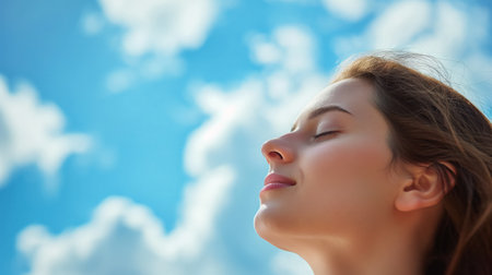 A serene woman stands with her eyes closed, head tilted back, and face bathed in light against a backdrop of a clear blue sky with fluffy clouds.の素材