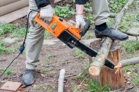 A man is sawing a log with an electric saw. Orange electro saw. Men's hands hold an electric saw, sawing a tree trunk.の写真素材