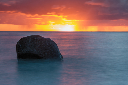 Sunrise at the Baltic sea with red horizon and blue water. In the foreground is a big stone in the waterの写真素材