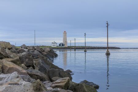 Lighthouse and uninhabited houses in the background. Lateral power pylons in the water. Stone coastline of the Grotta island near Reykjavik with clouds in the skyの写真素材