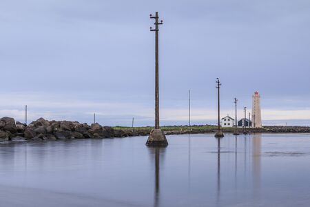 Stone coastline of the Grotta island near Reykjavik with clouds. Blue sky with power pylons in the water at the lighthouse and uninhabited houses. Peninsula with bird sanctuaryの写真素材