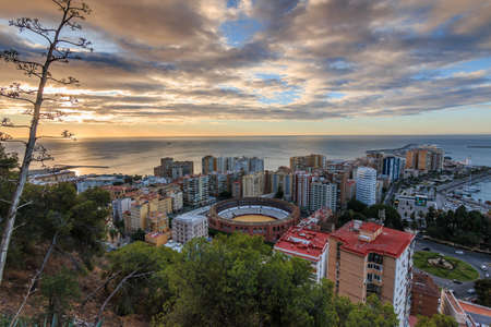 Spanish coast in Andalusia in the morning. View over the city of Malaga at sunrise with clouds and an orange horizon. City view on the Costa del Sol from park with trees and bushesの写真素材
