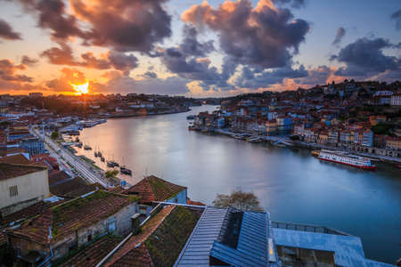 Sunset over the rooftops of the city of Porto. River Douro in the Portuguese coastal city. Clouds and sun rays in the evening. City view from an elevated position with harbor, river boats and hillsの写真素材