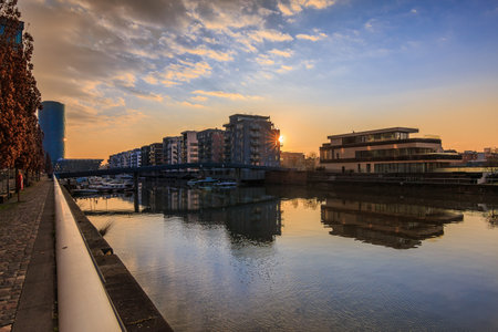 Residential area on the canal from the Main in Frankfurt at sunrise. Water front with reflection. Blue sky and clouds. Shore in the harbor and buildings with trees and brown leavesの写真素材