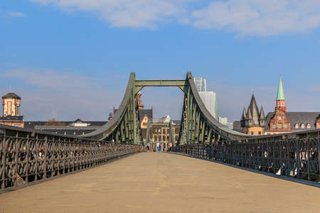 Pedestrian bridge over the river Main in Frankfurt. Iron bridge during the day with few people. Houses of the old town in the background with sunshine and few cloudsの写真素材