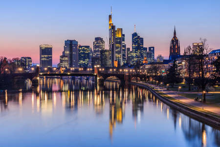 Frankfurt city skyline in the evening after sunset. Illuminated buildings of high-rise buildings from the business and financial center. Bridge over the river Main with street lightsの写真素材