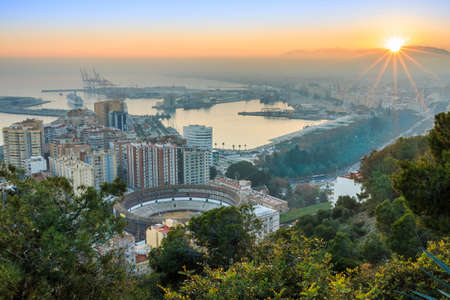 Evening sun over the Spanish city of Malaga. Sunset on the Spanish Costa del Sol with a panoramic view of the harbor, houses, trees, bullring with illuminated street lamp and blue skyの写真素材