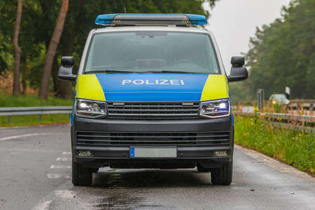 Front view of a police car on the highway with the lights on. Vehicle from the state of Brandenburg in a parking lot. Asphalt roadway of an emergency stop with guard rails and treesの写真素材