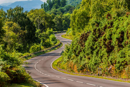 A narrow, winding road in Scotland along Loch Ness. Trees and bushes next to the road in summer in sunshine. Traffic signs and crash barriers with lane markings on the roadwayの写真素材