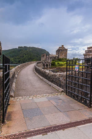 Historic stone bridge to Eilean Donan Castle during low tide. Open black gate to the bridge. Pedestrian bridge with three archways. Cloudy sky and hills in the background in summerの写真素材