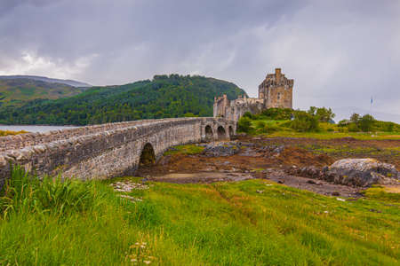 Eilean Donan Castle in Scotland with the historic stone bridge at low tide. Island with castle, hill and lake when the water is low. Bushes, grass and stones in the foregroundの写真素材