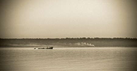 An old style photo with noise, scratches and vignette of an old rusty ship wreck in sea in front of a land with smoking bonfires の写真素材