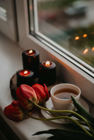 A cup of tea, a bouquet of red tulips and candles on the windowsill.の写真素材