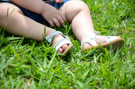 A girl with her pale-skinned legs, sitting on the green grass of the garden, feet wearing white and blue sandals, jeans shorts, little hand and little fingers playing in the grass.の写真素材