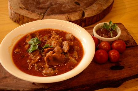 On a wooden board, a small white bowl, chicken gizzards in sauce, cherry tomatoes, basil and a...
