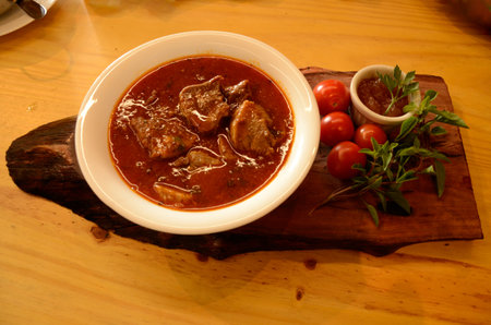 On a wooden board, a small white bowl with beef tongue in sauce, cherry tomatoes, basil and a...
