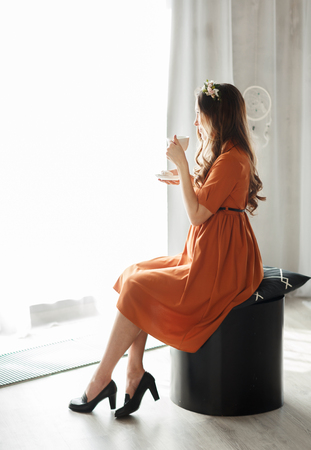 Portrait of happy pregnant woman in a dress with cup drinking tea looking through window at home. The concept of rest and pregnancyの写真素材