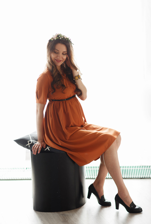 Beautiful pregnant mother in red dress posing at decorated studio, smiling at camera. Mom standing near window.の写真素材