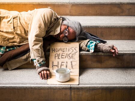 A poor homeless man sleep on a ladder with a donation billの写真素材